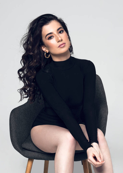 Front view of a sensual young woman with hairstyle wear black bodysuit sitting on chair, looking at camera, on a grey background. Vertical view. - Foto, afbeelding