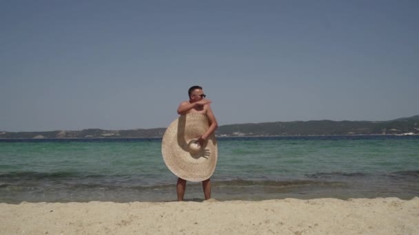 Larga oportunidad de hombre gordita divirtiéndose en la playa. Hombre en gafas de sol con sombrero de verano grande jugando cerca del agua, mirando a su alrededor - Imágenes, Vídeo