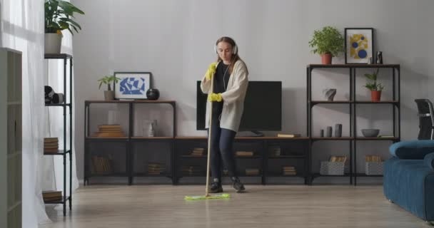 fröhliche Frau wäscht Boden im Wohnzimmer, tanzt mit Wischmopp und singt, hört Musik über Kopfhörer - Filmmaterial, Video