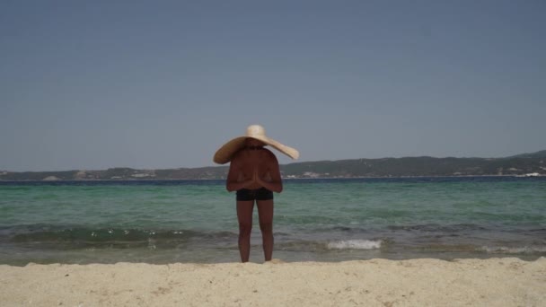 Un uomo che si diverte in spiaggia. Uomo in occhiali da sole con un grande cappello estivo che gioca vicino all'acqua, scherza e posa. - Filmati, video