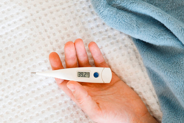Großaufnahme eines Mannes, der ein digitales Thermometer in der Hand hält, auf dem Bett liegend, drinnen. Hohe Körpertemperatur, Kälte, Grippeepidemie - Foto, Bild