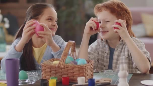 Портрет милого мальчика и девочки, сидящих дома, держащих окрашенные пасхальные яйца перед глазами, позирующих перед камерой и счастливо улыбающихся - Кадры, видео