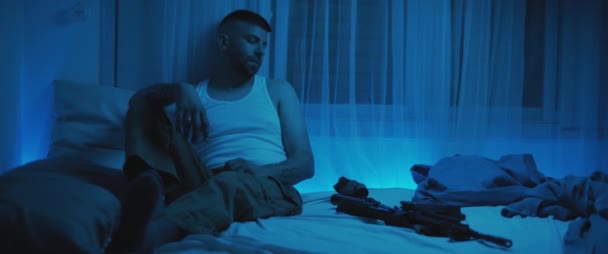 Ένας στρατιώτης που κάθεται σε μια σκοτεινή κρεβατοκάμαρα με ένα τουφέκι ξαπλωμένο στο κρεβάτι, ενώ αντιμετωπίζει μετα-τραύμα. Άγχος, κατάθλιψη, έννοια ψυχικής υγείας. Σε πραγματικό χρόνο, μπλε φωτισμός. - Πλάνα, βίντεο
