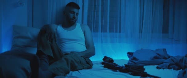 Ένας στρατιώτης κάθεται σε μια σκοτεινή κρεβατοκάμαρα με ένα τουφέκι ξαπλωμένο στο κρεβάτι, ενώ ασχολείται με μετατραυματικό στρες. Άγχος, κατάθλιψη, έννοια ψυχικής υγείας. Σε πραγματικό χρόνο, μπλε φωτισμός. - Πλάνα, βίντεο