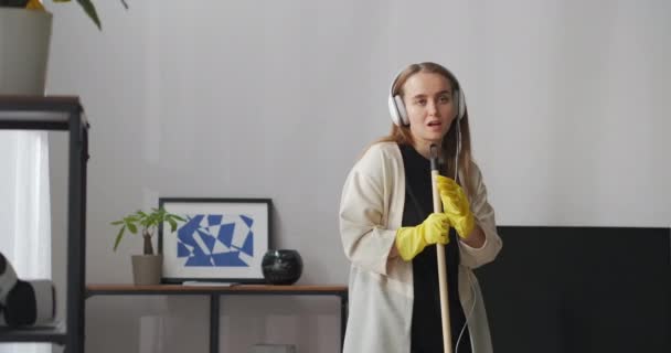 pretty young woman is listening to song by headphones and singing to mop during clean-up at home, medium portrait shot - Footage, Video