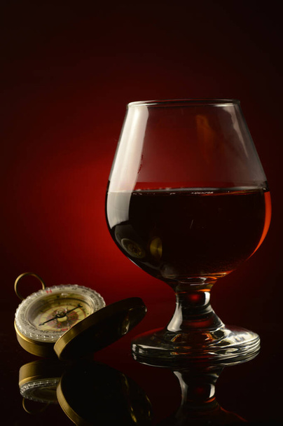 A vertical composition of a glass of Cognac and an old style compass over a dark atmospheric background. - Photo, image