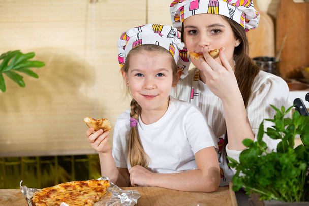 The five-year-old daughter sat down on her mothers lap and together they eat pizza made and baked by themselves. After working together in the kitchen, its time to eat together. - Foto, Bild