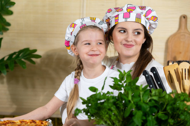 Porträt einer lächelnden fünfjährigen Tochter und Mutter mit geschwollenen Wangen, die eine frische Pizza essen. Nach der gemeinsamen Arbeit in der Küche ist es Zeit, gemeinsam zu essen. - Foto, Bild