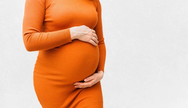 Pregnant woman in tight orange dress holds hands on belly on white background. Pregnancy and maternity concept. Close-up, copy space - Photo, image