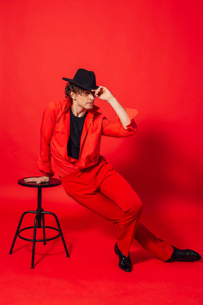 Portrait of a tall handsome man dressed in red shirt, red trousers and black hat posing with chair on the red background - Fotografie, Obrázek