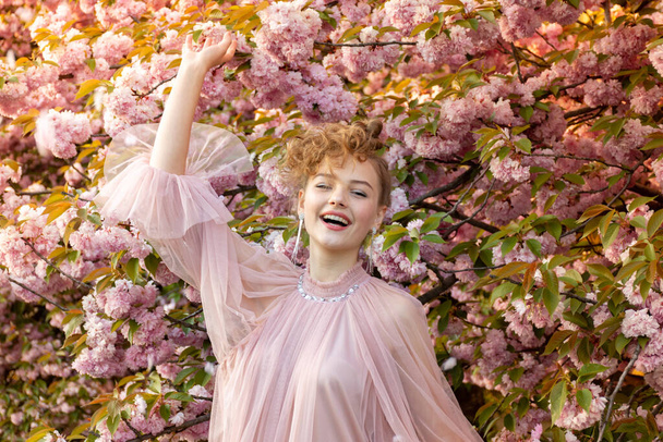 Very joyful young girl in a pink dress on a background of flowering sakura trees. Sincere emotions, laughter - Фото, зображення