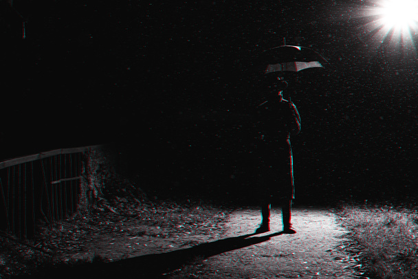dark silhouette of a man in a raincoat and hat under an umbrella on the street in the rain - Photo, Image
