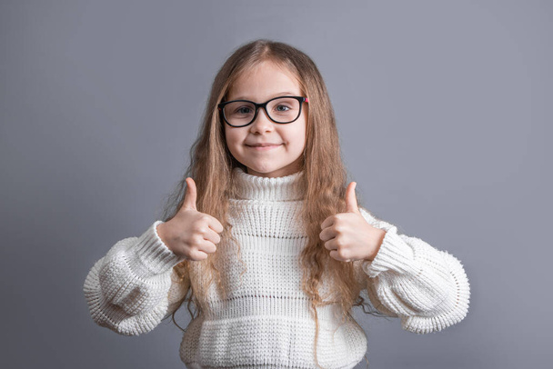Portret van een jong aantrekkelijk klein meisje met blond lang golvend haar in een witte trui die glimlacht toont duimen omhoog, als zingen op een grijze studioachtergrond. Plaats voor tekst. Kopieerruimte - Foto, afbeelding