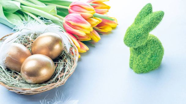 Egg color. Happy Easter decoration: golden colour eggs in basket with spring tulips, white feathers on pastel blue background. Foil minimalist egg design, modern template - Photo, Image