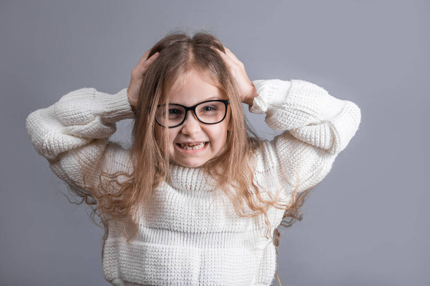 Portrait of a young attractive little girl with hands on head in a white sweater annoyed irritated pulling out her hair for nervous disagreement on a gray studio background. - Foto, afbeelding