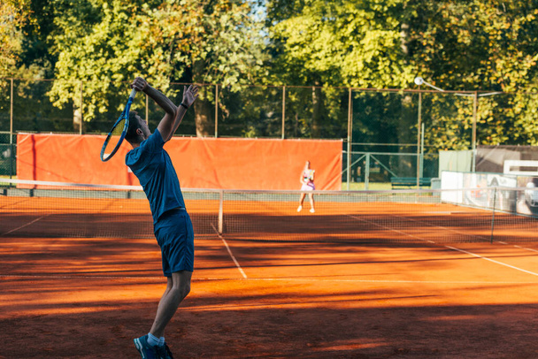 Visão traseira de um jogador de tênis pronto para servir em uma quadra de barro usando roupas esportivas azuis - Foto, Imagem