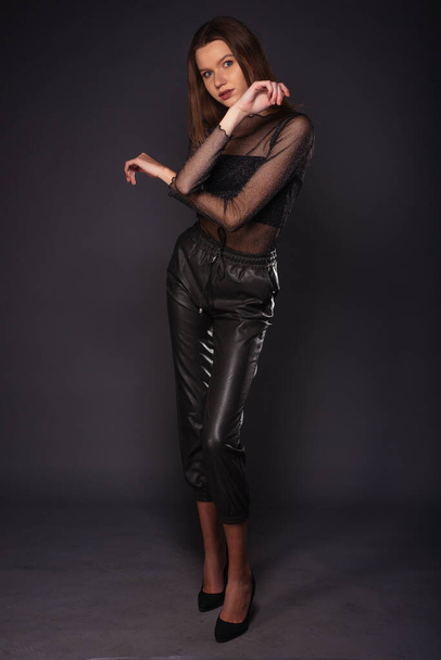 Ganztägiges Modeporträt einer attraktiven Frau in Lederhose und durchsichtigem Hemd, während sie auf dunklem Hintergrund posiert - Foto, Bild
