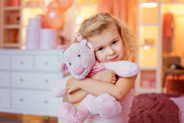 Una bambina bionda dai capelli ricci si siede sul letto e abbraccia un orsacchiotto rosa. - Foto, immagini