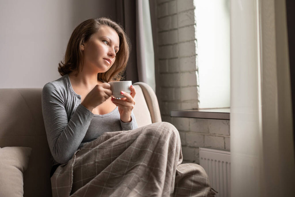 Portrait of beautiful caucasian young woman sitting on sofa covered blanket, drinking hot coffee from cup and looking at window. - Foto, imagen