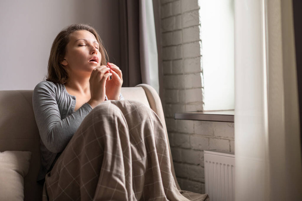 Sick brunette woman sneezing sitting on the couch in the living room near window - Foto, imagen