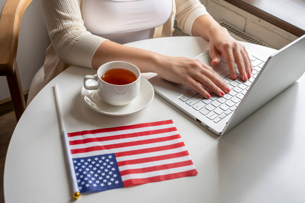 Lonely woman freelancer with flag of America enjoying having breakfast with cup of coffee working on laptop sitting near window in cafe at morning.  - Foto, Bild
