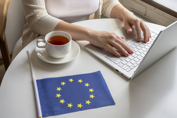 Lonely woman freelancer with flag of European Union enjoying having breakfast with cup of coffee working on laptop sitting near window in cafe at morning.  - Foto, Bild