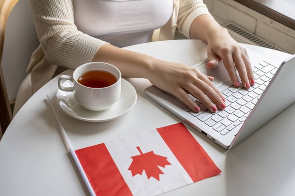 Одинокая женщина-фрилансер с флагом Канады наслаждается завтраком с чашкой кофе, работая на ноутбуке, сидя утром у окна в кафе.  - Фото, изображение