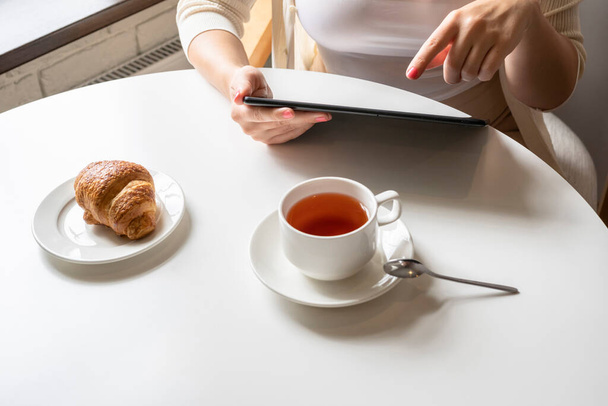 Lonely woman freelancer enjoying having breakfast with cup of coffee working on tablet sitting near window in cafe at morning. View on bust of female - 写真・画像