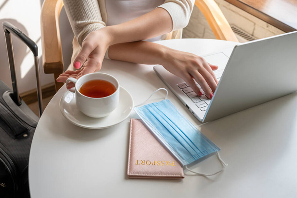 Woman tourist with cup of coffee and suitcase putting medical protective face mask and passport at the table sitting near window in cafe in airport using laptop. Travel concept during coronavirus - Foto, Imagen