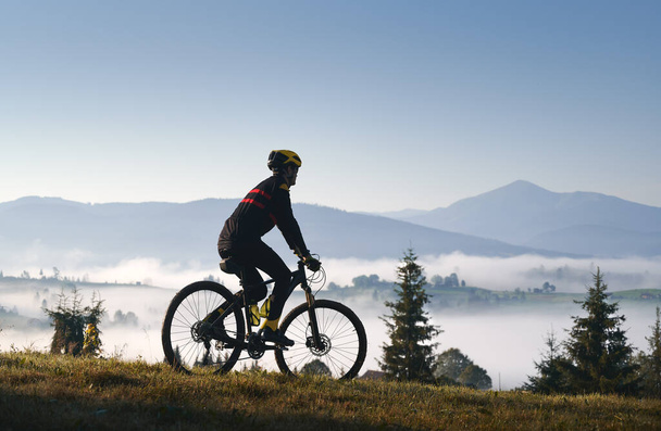 Egy férfi kerékpáros sziluettje biciklizős öltönyben biciklivel a füves dombon. Férfi kerékpáros élvezi a kilátást a fenséges hegyek kerékpározás közben. A sport, a kerékpározás és a természet fogalma. - Fotó, kép