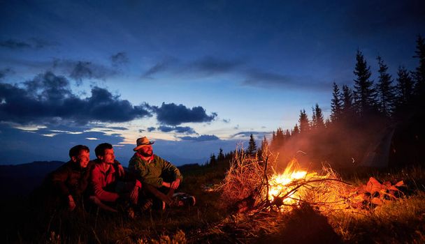 Summer recreation three tourists in the mountain camping with bonfire. Happy guys funny watching how burning dry brushwood on the backgraund of dusk sky. - Photo, Image