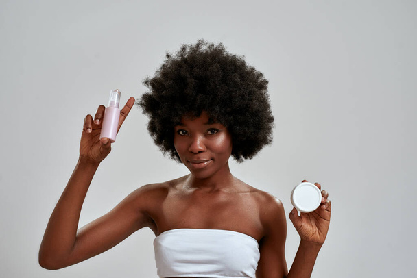 Attractive young african american woman with afro hair smiling at camera, choosing between two beauty products while standing isolated over gray background - Fotoğraf, Görsel
