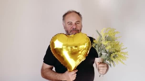 Handsom mature bearded man smiling and giving mimosa flower and heart ballon to viewer. Man with bouquet.Holiday concept - Footage, Video