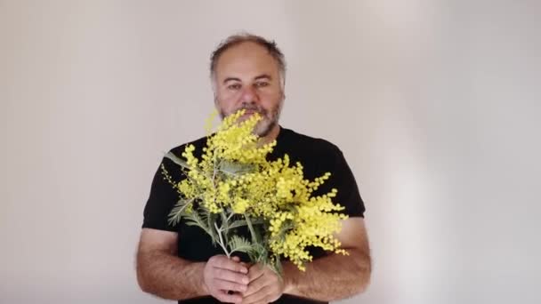 Handsom homme barbu mature souriant et donnant fleur de mimosa au spectateur. Homme avec bouquet.Concept de vacances. Mouvement lent. - Séquence, vidéo