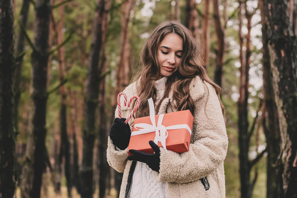Όμορφη νεαρή γυναίκα του μοντέλου εμφάνιση με μακριά σγουρά μαλλιά κρατά ένα χριστουγεννιάτικο δώρο και ένα γλειφιτζούρι και χαμόγελα, ενώ είναι σε ένα πευκοδάσος - Φωτογραφία, εικόνα