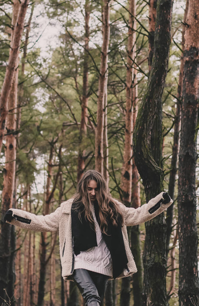 A slender woman of model appearance with long dark hair is wearing a fur coat, a sweater hat and leather pants and walks along a path in a pine forest in winter - Photo, Image