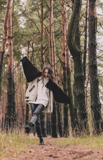 Стройная женщина образцового вида с длинными темными волосами одета в меховую шубу, свитер и кожаные брюки, держит шарф в руках и ходит по тропинке в сосновом лесу зимой - Фото, изображение