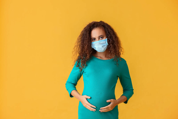 Gestresste schwangere Afroamerikanerin in medizinischer Maske auf farbigem Hintergrund - Foto, Bild