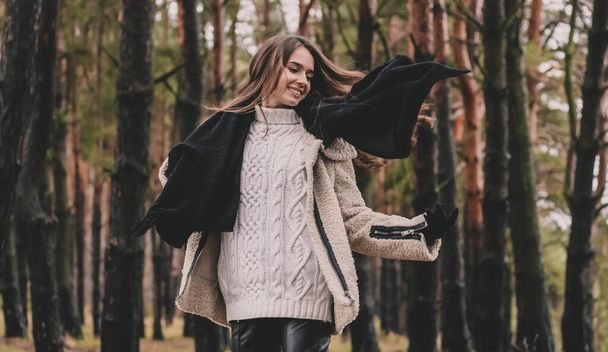 Kobieta wyglądająca jak modelka z długimi ciemnymi włosami, która lata na wietrze trzyma szalik w dłoniach i uśmiecha się podczas gdy w sosnowym lesie - Zdjęcie, obraz