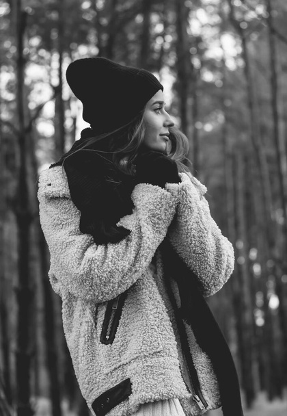 Een slanke vrouw met model uiterlijk met lang donker haar is gekleed in een bontjas, een trui hoed en leren broek loopt langs een pad tegen een achtergrond van een dennenbos in de winter - Foto, afbeelding
