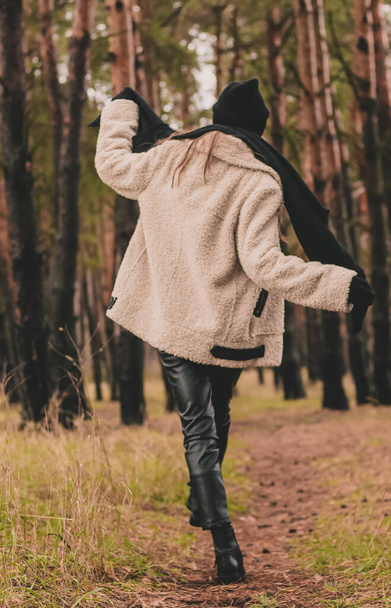 Μια λεπτή γυναίκα του μοντέλου εμφάνιση με μακριά σκούρα μαλλιά είναι ντυμένη με γούνα παλτό, ένα πουλόβερ καπέλο και δερμάτινο παντελόνι βόλτες κατά μήκος ενός μονοπατιού σε ένα φόντο ενός πευκοδάσους το χειμώνα - Φωτογραφία, εικόνα