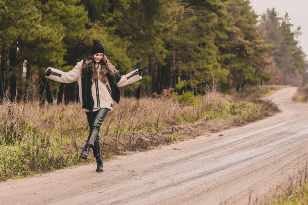 Uma mulher esbelta de aparência de modelo com cabelo escuro longo veste-se com um casaco de peles, um chapéu de suéter e calças de couro caminha ao longo de um caminho contra um contexto de uma floresta de pinheiro no inverno - Foto, Imagem