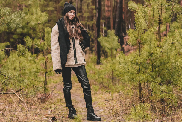 A beautiful young woman of model appearance with long curly hair in a sweater hat and a fur coat stands against the background of Christmas trees in a pine forest - Φωτογραφία, εικόνα