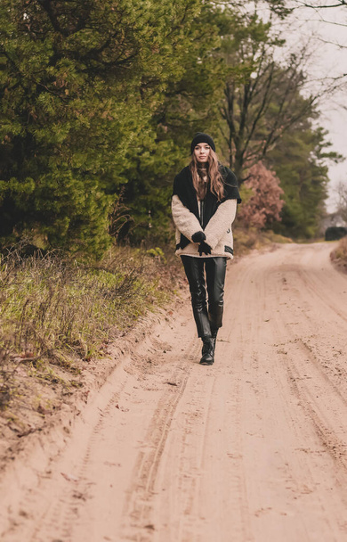 Una donna snella di aspetto modello con lunghi capelli scuri è vestita con una pelliccia, un cappello di maglione e pantaloni di pelle cammina lungo un percorso su uno sfondo di una pineta in inverno. - Foto, immagini