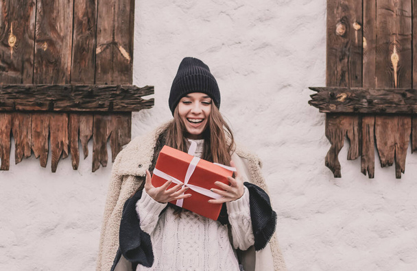Schöne junge Frau von modelliertem Aussehen mit langen lockigen Haaren, Pullovermütze und Pelzmantel, die ein Weihnachtsgeschenk in den Händen hält, vor weißem Hintergrund mit Baumfenstern - Foto, Bild