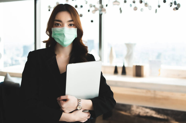 infection of the virus coronavirus.businesswoman entrepreneur wears face mask standing and holding laptop with confident, with colleague in background in meeting room at modern office.Health and teamwork concept - Foto, Imagem