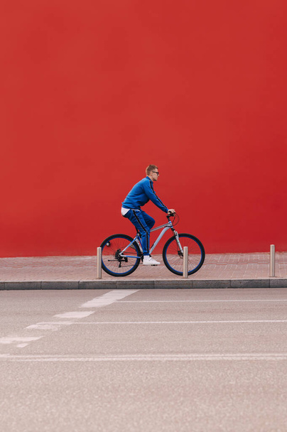 Man fietser in een blauw sportpak loopt op een fiets op straat op een achtergrond van een rode muur. Verticaal - Foto, afbeelding