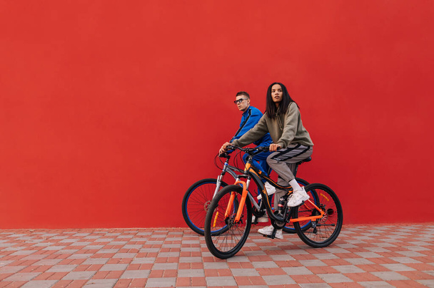 Kırmızı bir duvarın arka planında bisiklet süren şık bir erkek ve kadının fotoğrafı. Hafta sonları sokakta yürüyen şık spor yapan iki bisikletçi. - Fotoğraf, Görsel