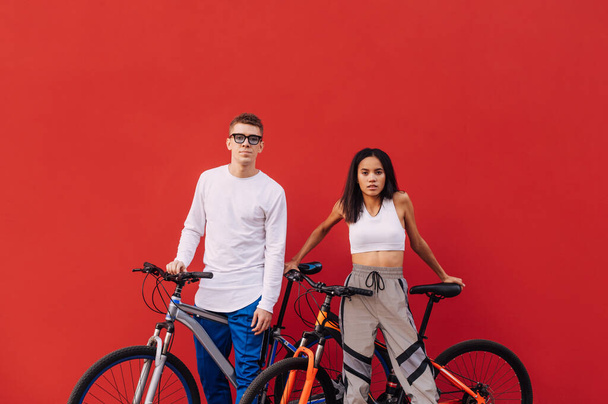 Gyönyörű pár kerékpárosok alkalmi ruhák állnak a piros háttérben kerékpárok és pózol a kamera. Stílusos ejtőernyős emberek egy kerékpáros edzés - Fotó, kép