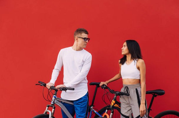 Hermosa pareja hombre y mujer en ropa casual con bicicletas aisladas en el fondo de la pared roja, descansando después de montar y sonriendo. - Foto, imagen